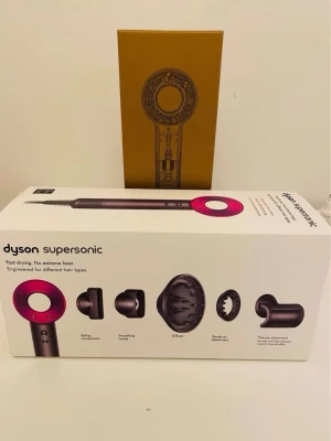 Dyson Supersonic hårfön med tillbehör - Säljer en Dyson Supersonic hårfön i metallicgrå och rosa med flera tillbehör: stylingmunstycke, diffuser, gentle air attachment och flyaway attachment. Kommer med originalkartong och manual. Perfekt för dig som vill ha snabb och skonsam hårtorkning med modern design.