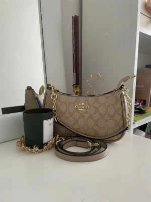 Beige axelväska från Coach - (priset går att diskuteras) Säljer en beige axelväska från Coach med klassiskt monogrammönster och guldfärgade detaljer. Väskan har avtagbar axelrem och kedjedetalj, samt Coach-tag i läder. Perfekt storlek för det viktigaste och riktigt snygg till alla outfits.