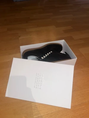 Svarta Maison Margiela sneakers - Maison Margiela sneakers i svart skinn med vita snören och vit insida. Klassisk låg modell med gummisula i beige och diskreta sömdetaljer. Snygg minimalistisk design som passar till det mesta. Kommer i originalkartong.