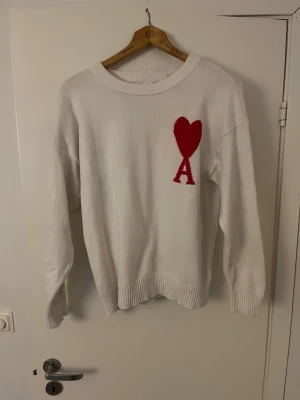 Vit stickad tröja från Ami - Vit stickad tröja från Ami med rund hals och ribbade muddar. På bröstet finns ett stort rött hjärta och bokstaven A som en snygg detalj. Tröjan har lång ärm och en normal passform, perfekt för dig som gillar stilrena plagg med en twist.