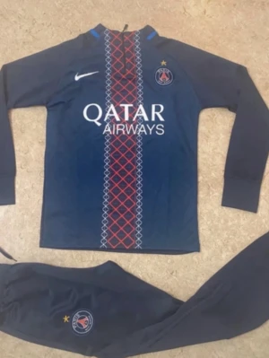 PSG träningsset i marinblått - Säljer ett PSG träningsset från Nike i marinblått med långärmad tröja och matchande byxor. Tröjan har rött och vitt mönster framtill, PSG-logga och Nike swoosh. Byxorna har PSG-logga och resår i midjan. Perfekt för fotbollsträning eller chill.
