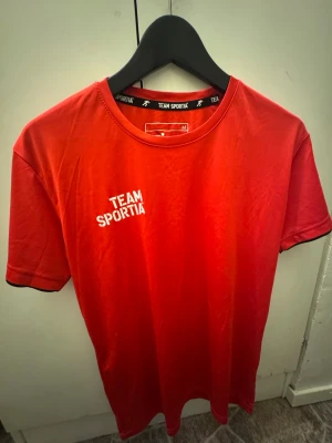 Tränings tröja  - En röd t-shirt från Team Sportia med texten 'MATCHENS LIRARE' på ryggen, inte några skor.