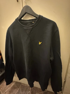 Svart sweatshirt från Lyle & Scott - Svart sweatshirt från Lyle & Scott med klassisk rund hals och ribbade muddar. Tröjan har ett broderat gult fågelmärke på bröstet och är tillverkad i mjuk bomull. Perfekt för chill dagar och enkel att matcha med jeans eller joggers.