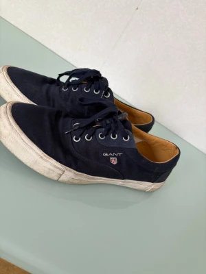 Marinblå sneakers från Gant - Snygga marinblå sneakers från Gant med klassisk design, vita gummisulor och snörning. Ovandelen är i bomullstyg och insidan har en beige läderdetalj. Gant-logga på sidan och bak på hälen. Perfekta för en avslappnad och stilren look.