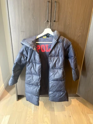 Mörkblå lång pufferjacka från Polo - Säljer en lång, mörkblå pufferjacka från Polo med huva och stora röda bokstäver på insidan. Jackan har dragkedja och tryckknappar framtill, samt quiltad design som håller värmen. Perfekt för kalla dagar och riktigt snygg till vardagslooken.