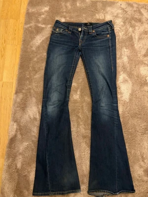 True Religion Joey Low Rise Flare Jeans - Säljer ett par mörkblå True Religion Joey jeans med låg midja och flare-ben. Klassiska kontrastsömmar och stora fickor bak med ikonisk broderad logga. Tillverkade i stretchig bomullsdenim för skön passform. Perfekta för dig som gillar en retro och statement look. Tyvär saknar en ficka en knapp och jeansen har även ett hål vilket syns tydligt på bilden. 