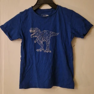 Blå t-shirt med dinosaurie från Carry Kids - Blå t-shirt från Carry Kids i storlek 122 med ett coolt vitt dinosaurietryck på bröstet. Klassisk rund hals och korta ärmar. Hund finns i hemmet. Kolla in mina andra annonser för samfrakt! 
