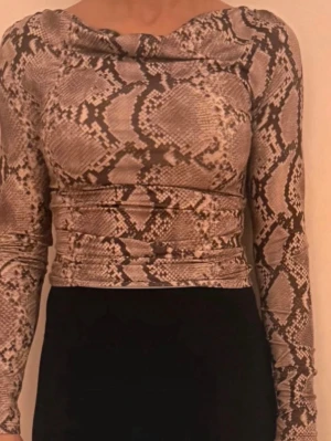 Brun långärmad topp med ormmönster - Trendig långärmad topp med ormmönster i bruna och beige toner. Toppen har en åtsittande passform och en snygg draperad halsringning. Perfekt att styla med höga byxor eller kjol för en cool look.