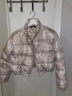Beige croppad pufferjacka - Säljer en beige croppad pufferjacka från River Island med stora vita bokstäver som mönster. Jackan har dragkedja framtill, hög krage och långärmade puffiga ärmar. Perfekt för dig som vill ha en trendig och varm jacka med cool streetstil.