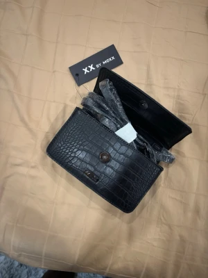 Svart axelväska från XX by Mexx - Snygg svart axelväska från XX by Mexx i croco-mönstrat skinnimitation. Väskan har ett stilrent lock med magnetknäppning och justerbar axelrem. Perfekt storlek för det viktigaste och ger en trendig touch till din outfit.