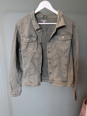 Olivgrön jeansjacka från Sincerely Loved - Snygg olivgrön jeansjacka från Sincerely Loved i klassisk modell med knappar framtill och två bröstfickor med lock. Jackan har lång ärm och en rak passform som funkar till många olika stilar. Perfekt att slänga på sig när det är lite kyligt ute.