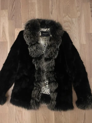 Svart pälsjacka från Hollies - Säljer en svart pälsjacka från Hollies med fluffig krage och muddar i grå päls. Jackan har ett snyggt mönstrat innerfoder och en klassisk rak passform. Perfekt för dig som vill ha en statement piece med lyxig känsla.