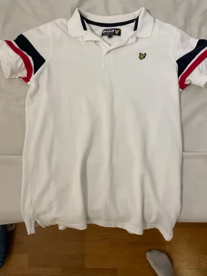 piké från Lyle & Scott - Säljer en vit pikétröja från Lyle & Scott med klassisk krage och knappar. Pågrund av att den blivit för liten. Storleken är 14/15 år men passar som en xs. Tröjan har marinblå och röda detaljer på ärmarna samt den ikoniska gula loggan på bröstet. Tillverkad i mjuk bomull och har en normal passform. Perfekt till sommaren!