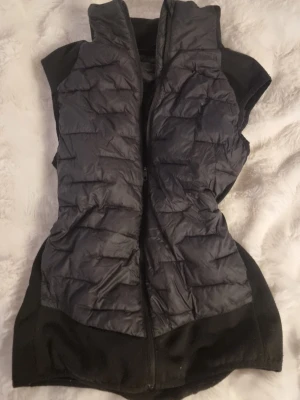 Svart dunväst från Everest, strl 38 - Svart dunväst från Everest med quiltad framsida och diskret logga på bröstet. Västen har hög krage som går att vinkla ner, fickor och dragkedja framtill. Materialet funkar för många vänder, även perfekt för lager på lager. Snygg och sportig look. Sidorna är i mjukare material för extra komfort, perfekt för att ha hoodie eller liknande under.