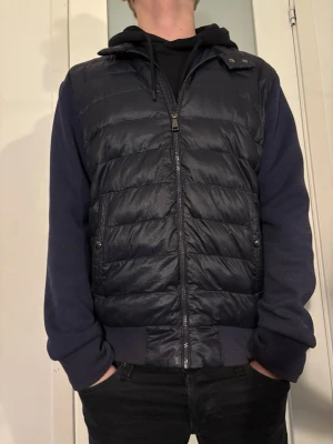 Mörkblå pufferjacka Polo Ralph Lauren - Säljer en mörkblå pufferjacka från Polo Ralph Lauren med quiltad front, mjuka ärmar och huva. Jackan har dragkedja framtill, två sidofickor med knapp och broderad logga på ärmen. Perfekt för dig som vill ha en stilren och sportig look. OBS liten defekt fram (Bild 6).