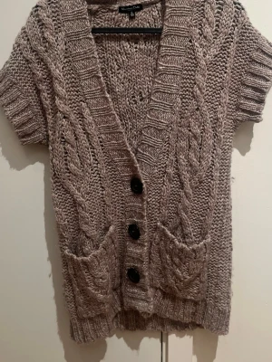 Beige stickad kofta Massimo Dutti - Kortärmad beige stickad kofta från Massimo Dutti i storlek S. Grovstickad med flätmönster, djup v-ringning och stora svarta knappar framtill. Två fickor fram och ribbade kanter. Materialet är 50% ull och 50% akryl.