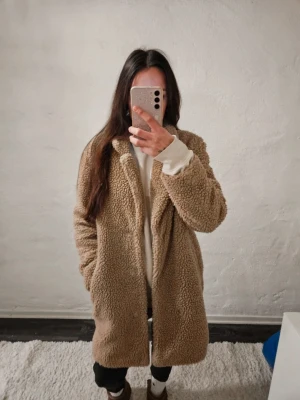 Beige teddyjacka med krage - Mysig beige teddyjacka med klassisk krage och knäppning framtill. Jackan är lång och har en oversized passform. Fodrad insida och stora fickor på sidorna.