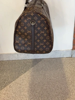 Louis Vuitton brun resväska monogram - Stor och rymlig resväska från Louis Vuitton i klassiskt brunt monogrammönster. Väskan har två robusta handtag, detaljer i mörkbrunt och dragkedja upptill. Tillverkad i slitstarkt skinn med ikoniska LV-loggor och blommor över hela ytan.