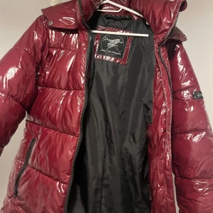 Vinröd pufferjacka från Save The Duck - Säljer en glansig vinröd pufferjacka från Save The Duck med svart innerfoder och dragkedja. Jackan har , två sidofickor med dragkedja och en innerficka. Perfekt för kalla dagar och riktigt snygg färg!