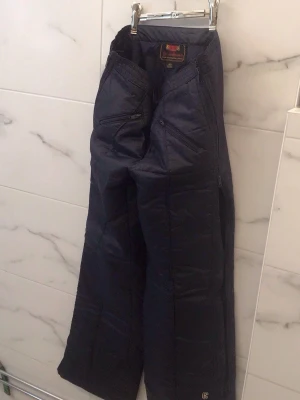 Svarta täckbyxor från Åre Skidbyxor - Säljer ett par svarta täckbyxor från Åre Skidbyxor med dragkedjor längs benen och flera fickor med dragkedja. Byxorna har elastisk midja och är perfekta för kalla dagar i backen. Materialet är slitstarkt och isolerande för att hålla dig varm.