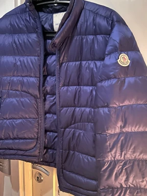 Mörkblå pufferjacka från Moncler - Säljer en mörkblå pufferjacka från Moncler med klassisk quiltad design och hög krage. Jackan har dragkedja framtill, sidofickor med dragkedja och Moncler-logga på ärmen. Perfekt för kalla dagar och riktigt snygg till streetwear.