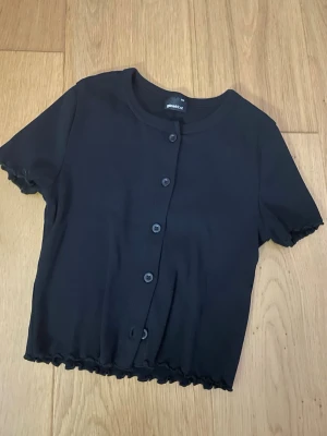 Svart kortärmad kofta från Gina Tricot - Säljer en svart kortärmad kofta från Gina Tricot. Modellen är croppad med knappar framtill och har vågiga kanter vid ärmslut och nederkant. Perfekt till sommaren och enkel att styla med olika outfits.