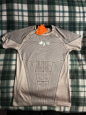 YoungLA Batman  - Säljer en grå kortärmad compression från YoungLA med Batman logga i silver på bröstet. Tröjan har ett unikt hexagonmönster över hela plagget och är tillverkad i ett stretchigt syntetmaterial som passar perfekt till träning. Tight passform för en sportig look.