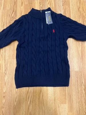 Marinblå stickad tröja Polo Ralph Lauren - Snygg marinblå stickad tröja från Polo Ralph Lauren med kabelstickat mönster och röd broderad logga på bröstet. Tröjan har långärmad design, ribbad krage och half zip-dragkedja vid halsen. Perfekt för dig som gillar klassisk stil med en modern touch.