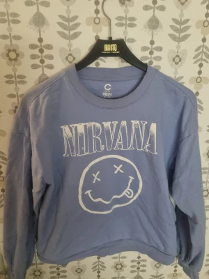 Ljusblå Nirvana sweatshirt - Säljer en ljusblå sweatshirt med Nirvana-tryck i vitt framtill. Tröjan har rund hals, långa ärmar och är gjord i mjuk bomull. Perfekt för dig som gillar bandtröjor och vill ha en avslappnad stil.