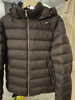 Mörkblå black label Moncler pufferjacka  - Säljer en svart/mörkblå pufferjacka från Moncler med huva och snygga detaljer som dragkedja och snörning vid kragen. Jackan har ett stilrent Moncler-märke på ärmen och är tillverkad i lätt och skyddande polyester. Perfekt för kalla dagar och har en modern. Den är storlek M men passar till S