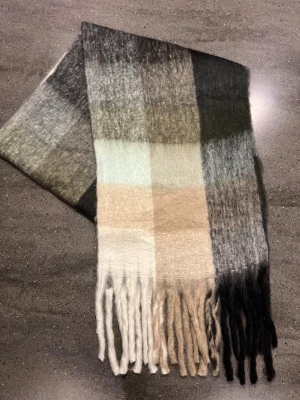 Halsduk - Snygg och mjuk halsduk ( som liknar acne studios) med rutigt mönster i svart, grått, beige och vitt. Den är lång och har tjocka fransar i ändarna. Perfekt att svepa runt halsen för extra värme och stil. Materialet känns fluffigt och lyxigt, passar dig som gillar neutrala färger.