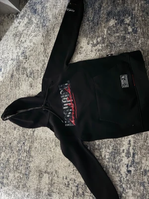 Svart hoodie från Hoodrich XS - Svart hoodie från Hoodrich i storlek XS med broderad logga i grått och rött på bröstet. Tröjan har huva med snörning, stor magficka och en streetwear-patch nedtill. Materialet är mjukt och känns som bomull, perfekt för chill dagar.