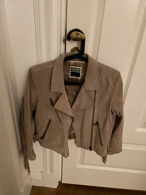 Beige mockajacka med fransar Weekday - Säljer en beige mockajacka från Weekday i storlek S. Jackan har coola fransar längs ryggen och ärmarna, flera sneda dragkedjor framtill och en croppad passform. Perfekt för dig som vill sticka ut med en unik look.