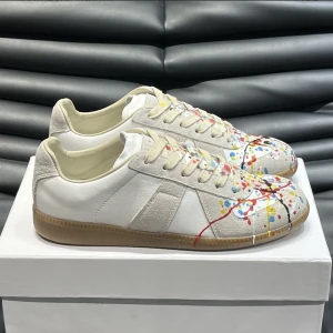 Vita Maison Margelas med färgstänk - Snygga vita sneakers med beige detaljer och färgstänk i rött, blått, gult och svart över tåpartiet. Skorna har snörning, platt sula och en mix av skinn och mocka som ger en cool och konstnärlig vibe. Perfekt för dig som vill sticka ut med din stil.