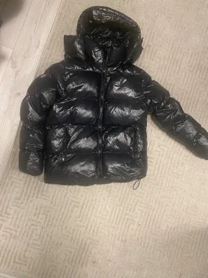 Svart pufferjacka från Everest - Säljer en svart pufferjacka från Everest med avtagbar huva och två fickor med dragkedja. Jackan har en glansig finish och är perfekt för kalla dagar. Stängs med dragkedja framtill och har en klassisk, puffig look. Org pris 1200