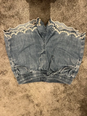 Blå denimshorts med spetskant - Säljer ett par ljusblå denimshorts med elastisk midja och snörning. Shortsen har en snygg volangkant upptill med vit spets och broderade detaljer som ger en cool och söt vibe. Perfekta för sommarens alla äventyr!