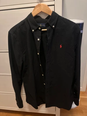 Svart skjorta från Ralph Lauren M - Svart skjorta från Ralph Lauren i storlek M. Klassisk button-down krage, vita knappar och rött broderat Polo-logo på bröstet. Skjortan har lång ärm och är gjord i mjuk bomull. Snygg och stilren modell som funkar till många olika looks.