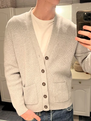 Grå cardigan  - Snygg ljusgrå cardigan med ribbad struktur och mörka knappar framtill. Modellen har två praktiska fickor och en djup v-ringning. Perfekt att slänga över en t-shirt för en chill look. Materialet är mjuk bomull och passformen är normal.