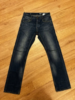 Mörkblå jeans från G-Star RAW - Snygga mörkblå jeans från G-Star RAW med klassisk femficksdesign och subtila slitningar på benen. Raka ben och kontrastsömmar ger en cool look. Perfekt för dig som gillar streetstyle och vill ha ett par jeans med lite edge.