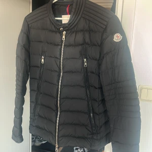 Svart Moncler pufferjacka herr äkta!! - Svart ny/vintage jacka från Moncler med quiltad design och coola detaljer som silverfärgade dragkedjor och ribbad krage. Jackan har lång ärm, två snedställda fickor med dragkedja och Moncler-logga på ärmen. Perfekt för dig som gillar stilren och sportig look. Väldigt unik jacka som är svår att fåtag på idag. Gott skick 