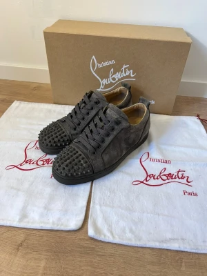 Christian Louboutin grå mocka sneakers - Säljer ett par grå sneakers från Christian Louboutin i mocka med coola nitar på tån och klassisk röd sula. Skorna har snörning och rund tå, samt en platt sula. Perfekt för dig som vill sticka ut med exklusiva detaljer och edgy vibe.