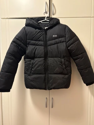 Svart pufferjacka från BOSS - Svart pufferjacka från BOSS med huva och snyggt broderad logga på bröstet. Jackan har dragkedja framtill, två fickor och quiltad design. Perfekt för kalla dagar och har en modern, loose passform som ger en chill vibe. Den är för barn i ungefär 9 till 13 åriga 
