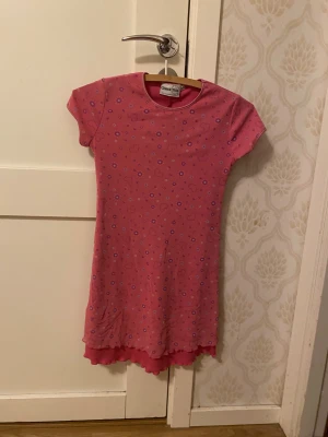 Rosa blommig klänning från Lindex - Supersöt rosa kortklänning från Lindex med små blommor och hjärtan i lila och vitt. Klänningen har korta ärmar, rund halsringning och dubbla volangkanter nertill. Perfekt för dig som gillar färg och mönster!