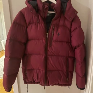 Vinröd pufferjacka från Polo Ralph Lauren - Säljer en vinröd pufferjacka från Polo Ralph Lauren med huva och flera dragkedjefickor. Jackan har en mörkblå insida, broderad logga på bröstet och kraftig vaddering som håller dig varm. Perfekt för kalla dagar och har en snygg, sportig look.