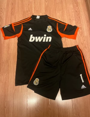 Real Madrid Casillas målvaktströja Adidas - Säljer en svart och orange Real Madrid målvaktströja med Casillas och nummer 1 på ryggen. Adidas-logga, bwin-tryck och klubbmärke på bröstet. LFP-patch på ärmen. Matchande shorts med klubbmärke och Adidas-logga. Tillverkad i polyester, kortärmad modell.
