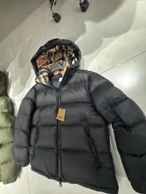 Svart Burberry pufferjacka med huva - Säljer en svart pufferjacka från Burberry med ikoniskt rutigt foder i huvan. Jackan har två stora fickor med dragkedja, quiltad design och en rejäl huva. Skriv din längd och vikt tack har alla storlekar. 2 färger Svart och Mörk Grön 