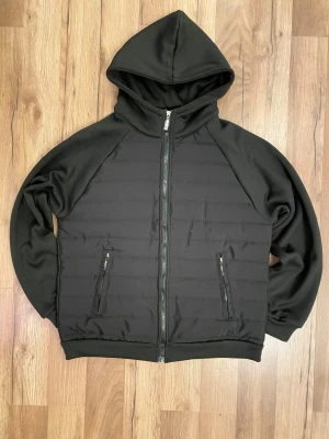 Cardigan - Svart cardigan med huva och hel dragkedja framtill. Jackan har två fickor med dragkedja och quiltad front samt insida. Ärmar och huva är i mjukare material, vilket ger en schysst kontrast. Perfekt för dig som gillar stilren och enkel look. 