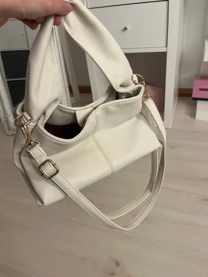 Vit axelväska i skinnimitation - Stilren vit axelväska med justerbar rem och snygga guldfärgade metalldetaljer. Väskan har en mjuk, fyrkantig form och är tillverkad i skinnimitation med lätt struktur. Perfekt storlek för att få plats med allt du behöver till vardagen.