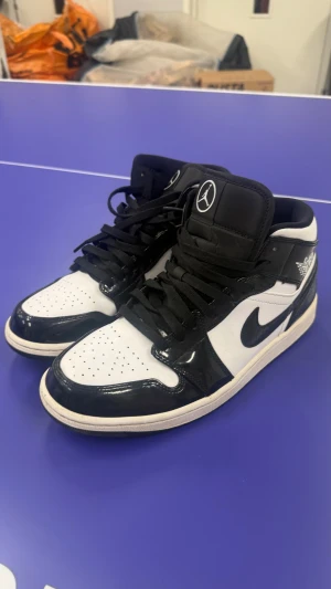 Nike Air Jordan 1 carbon mid  - Nike Air Jordan 1 sneakers i carbon och vitt med blankt, lackat skinn och klassisk Jordan-logga på sidan och plösen. Snygg kontrast mellan svart och vit, med svart snörning och rund tå. Sulan är vit med svart yttersula. Perfekt för dig som gillar streetstyle och ikoniska sneakers.