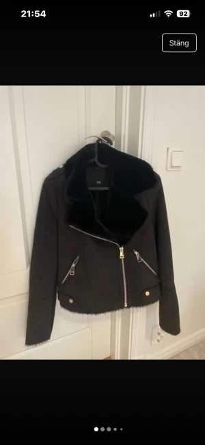 Svart pälsjacka med dragkedja - Svart pälsjacka med bred fluffig krage och snygga silvriga dragkedjor framtill och på fickorna. Jackan har guldknappar på axlarna och nertill, samt råa kanter för en cool look. 